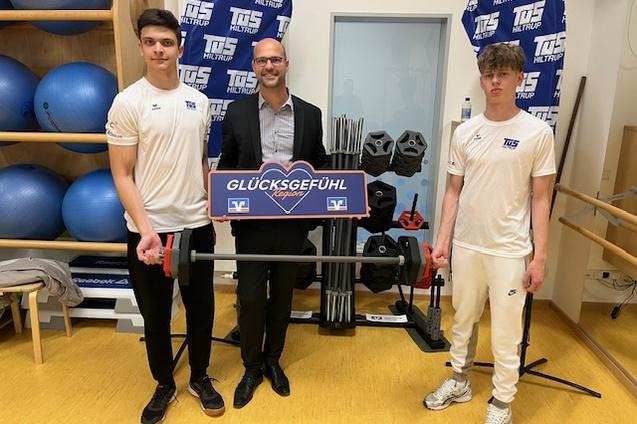 Drei Personen in Sportkleidung halten ein Schild mit der Aufschrift „GLÜCKSGEFÜHL“ vor Fitnessgeräten und Gewichten.