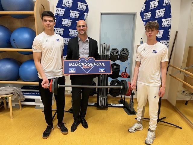 Drei Personen in Sportkleidung halten ein Schild mit der Aufschrift „GLÜCKSGEFÜHL“ vor Fitnessgeräten und Gewichten.