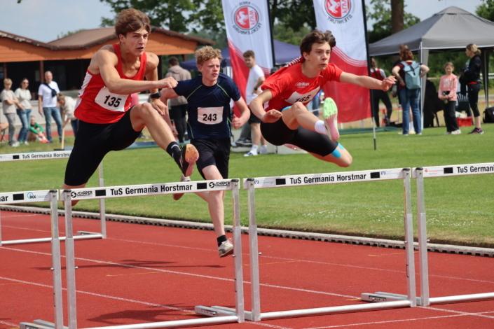 Drei Jugendliche laufen beim Hürdenlauf auf einer roten Laufbahn, zwei springen über die Hürden, Zuschauer im Hintergrund.