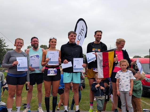 Sieben Läufer stehen auf dem Siegertreppchen mit Urkunden und einer Flagge, im Hintergrund ist eine Sportarena sichtbar.