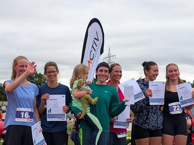 Gruppe von Frauen mit Urkunden nach einem Laufwettbewerb, eine hält ein Kind, im Hintergrund Event-Banner und Zuschauermenge.