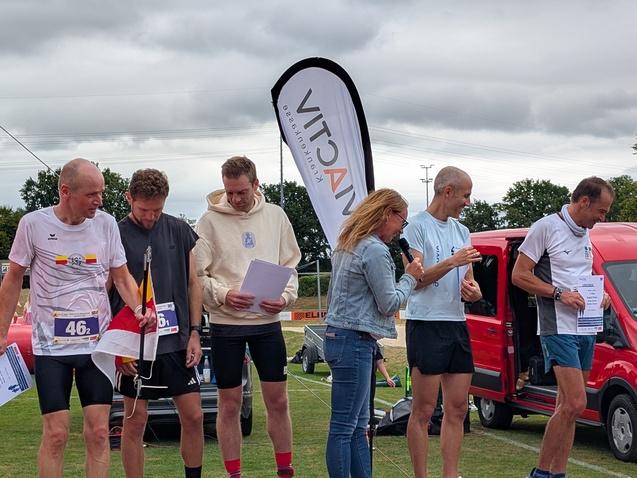 Sechs Teilnehmer stehen bei einer Siegerehrung, während eine Frau mit Mikrofon die Veranstaltung moderiert.