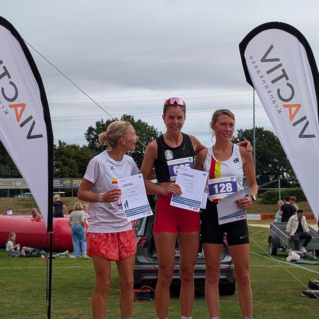 Drei Läuferinnen stehen auf dem Podest und halten ihre Urkunden, mit Flagge im Hintergrund.