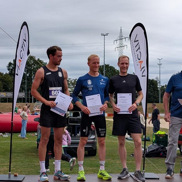 Drei männliche Läufer stehen auf einem Podium und halten Urkunden bei einer Sportveranstaltung im Freien.