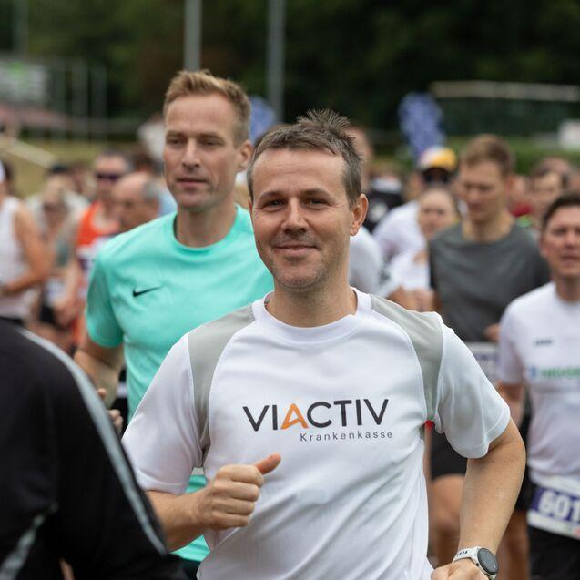 Gruppe von L&auml;ufern, die an einem Wettlauf teilnehmen, mit Fokus auf einen Mann in einem wei&szlig;en T-Shirt mit Logo.