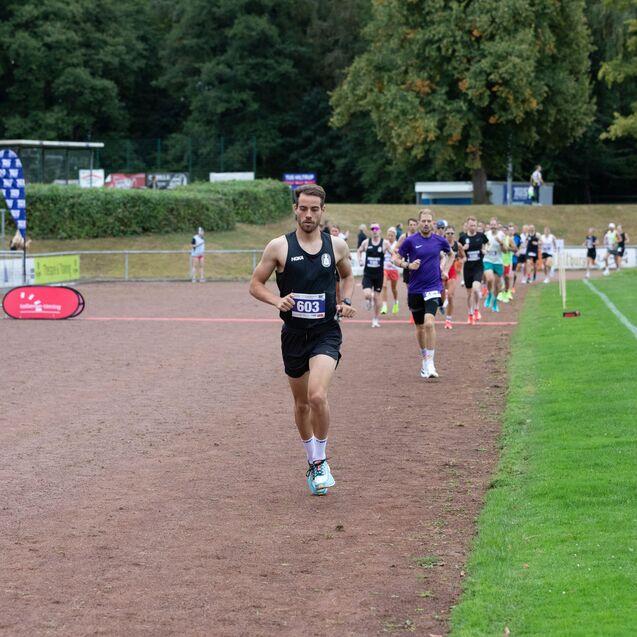 Ein L&auml;ufer mit Startnummer 603 joggt auf einer Laufbahn, w&auml;hrend weitere Teilnehmer im Hintergrund laufen.