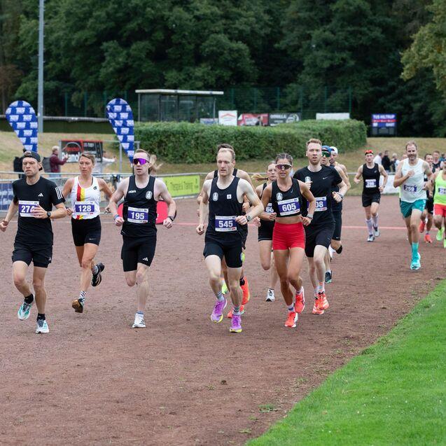 Gruppe von L&auml;ufern in Sportkleidung auf einer Laufbahn w&auml;hrend eines Wettkampfs, im Hintergrund Zuschauer und Werbebanner.