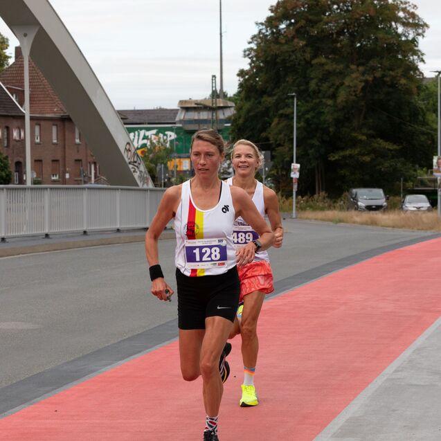 Zwei Frauen in Sportbekleidung laufen auf einer rot markierten Laufstrecke einer urbanen Umgebung.