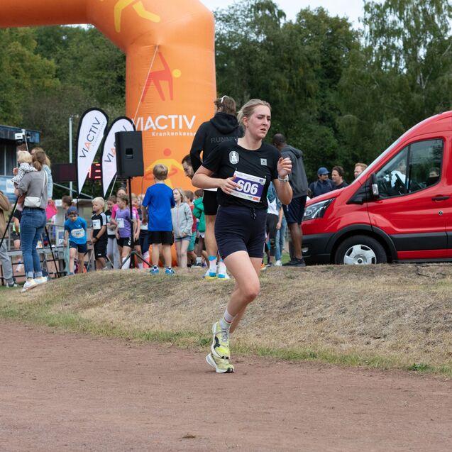L&auml;uferin mit Startnummer 506 beim Wettkampf, hinter ihr Kinder und ein rotes Fahrzeug auf der Sportanlage.