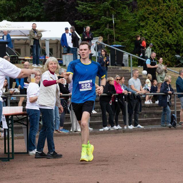 L&auml;ufer in blauer Sportbekleidung erh&auml;lt Wasser von Helfern bei einem Halbmarathon, Publikum beobachtet den Wettbewerb.
