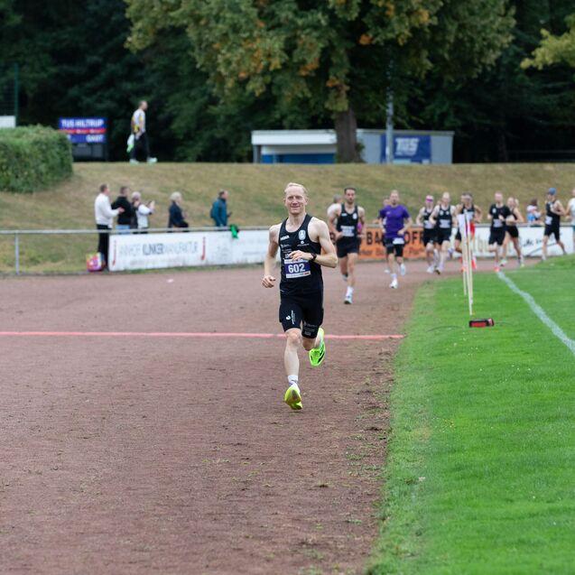 Läuft ein Sportler mit der Startnummer 602 auf einer Laufbahn, während Zuschauer und andere Läufer im Hintergrund sichtbar sind.