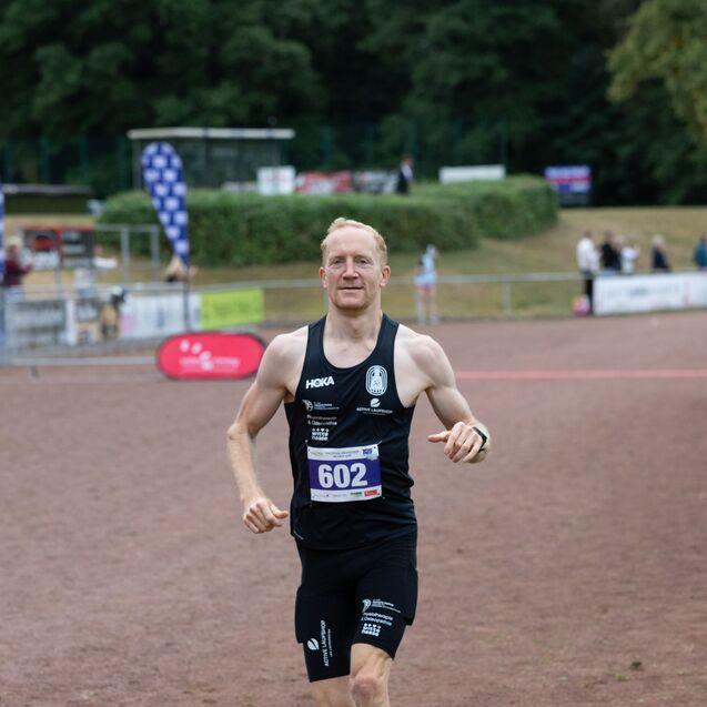 Läufer in Wettkampfkleidung überquert die Ziellinie auf einer Laufbahn, im Hintergrund Zuschauer und andere Teilnehmer.