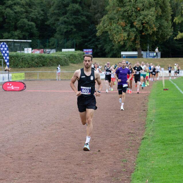 Ein Läufer in schwarzem Outfit mit Nummer 603 führt eine Gruppe von Athleten auf einem Außensportplatz an.