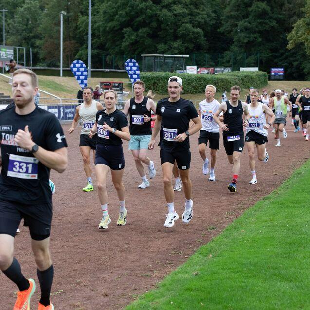 Eine Gruppe von Läufern bei einem Wettkampf auf einer Laufbahn, umgeben von Zuschauern und Sponsorenbanner.