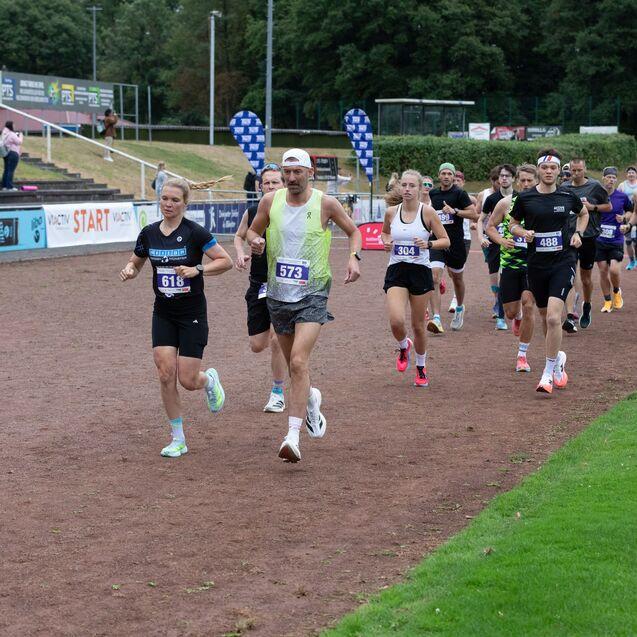 Gruppierter Lauf durch eine Sportanlage mit mehreren Teilnehmern in Sportkleidung und Laufnummern auf der Rennstrecke.
