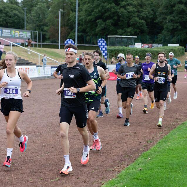 Gruppe von Läufern in Sportkleidung beim Wettlauf auf einer günen Laufbahn. Zuschauer stehen am Rand.