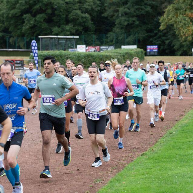Eine Gruppe von Läufern in Sportbekleidung läuft auf einer Laufbahn bei einem Event. Bäume und Werbebanner im Hintergrund.