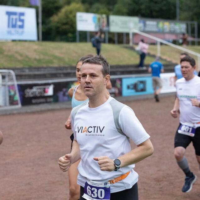 Läufer in Sportkleidung bei einem Wettlauf auf einem geschotterten Platz, im Hintergrund weitere Teilnehmer.