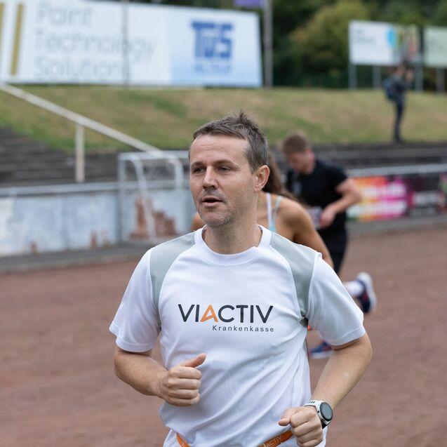 Läufer in einem weißen Shirt mit "VIACTIV" joggt auf einer Sportbahn, andere Teilnehmer sind im Hintergrund sichtbar.