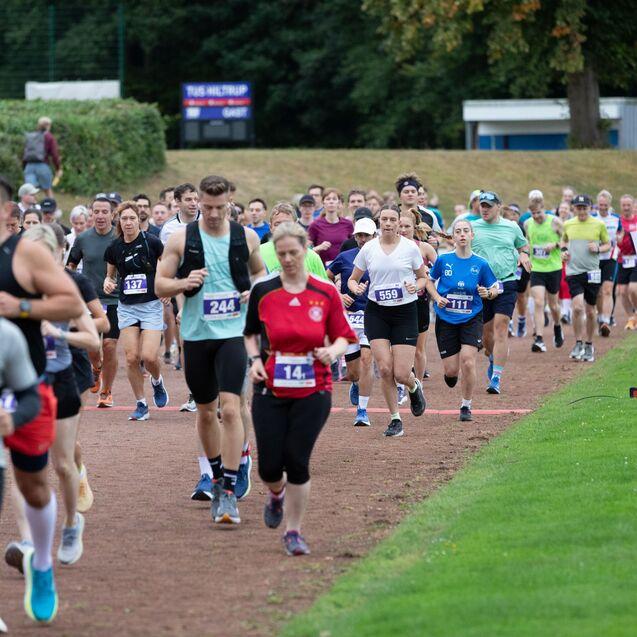Eine Gruppe von Läufern nimmt an einem Wettlauf auf einer Laufbahn teil, umgeben von Zuschauern und grüner Landschaft.