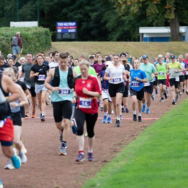 Eine Gruppe von L&auml;ufern nimmt an einem Wettkampf auf einer roten Laufbahn teil, umgeben von Rasen und B&auml;umen.