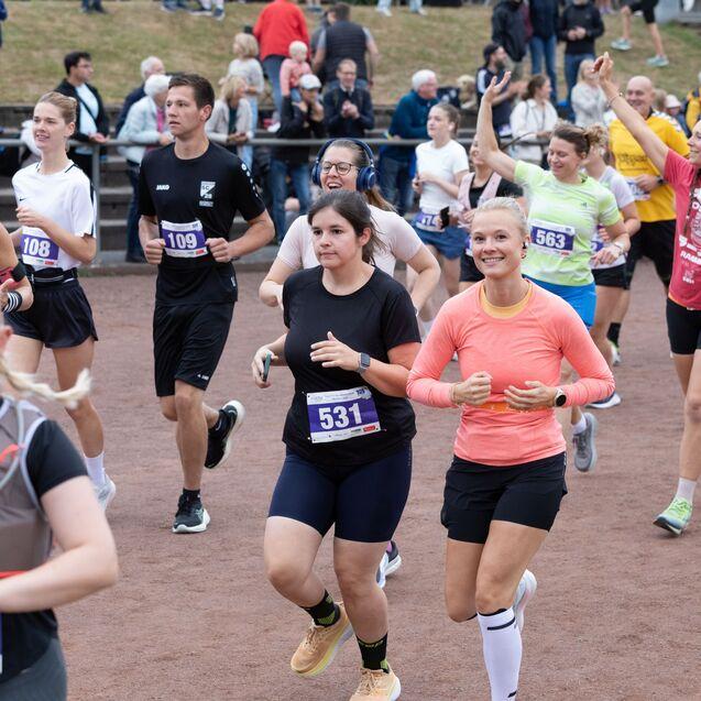 Gruppierung von L&auml;ufern in Sportbekleidung, die auf einem Laufplatz an einem Wettkampf teilnehmen. Zuschauer im Hintergrund.