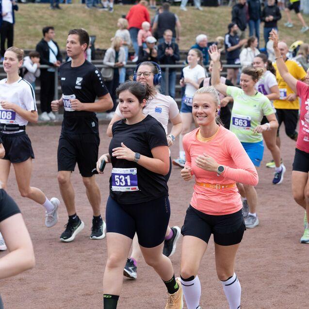 Läuferinnen und Läufer in Sportkleidung während eines Wettkampfs auf einer Sandbahn, Zuschauer im Hintergrund.