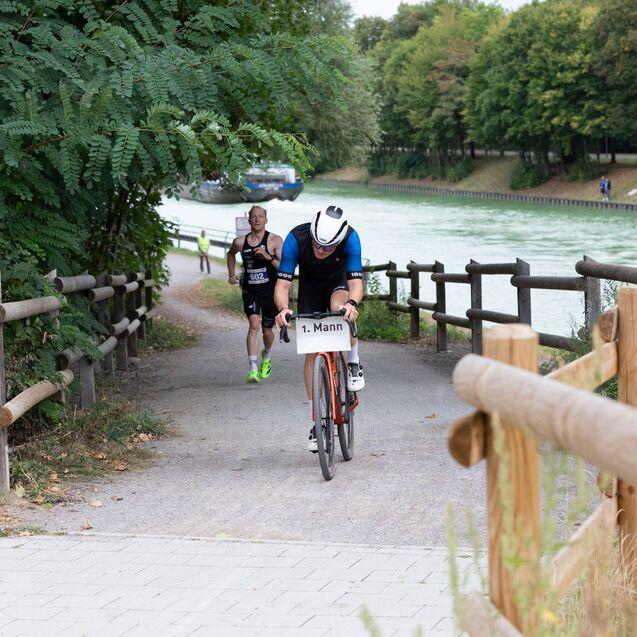 Ein Radfahrer im Sportoutfit f&auml;hrt auf einem Radweg, w&auml;hrend ein L&auml;ufer im Hintergrund l&auml;uft. B&auml;ume s&auml;umen den Weg.