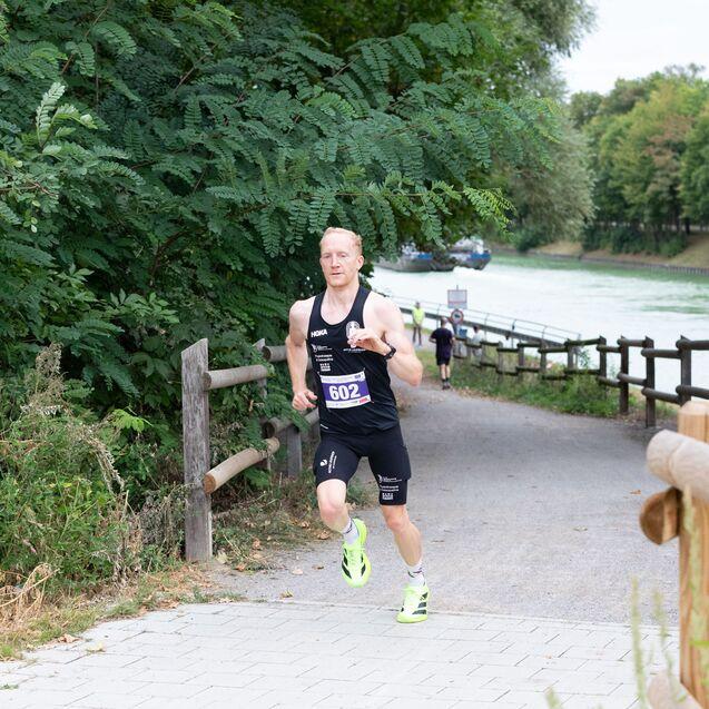 L&auml;ufer in Sportbekleidung sprintet auf einem Weg entlang eines Flusses, umgeben von B&auml;umen und gr&uuml;ner Vegetation.