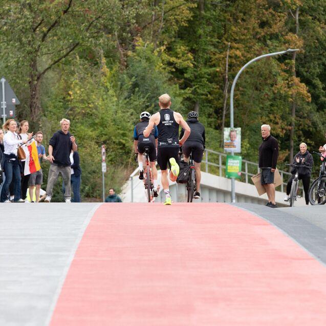 L&auml;ufer in Wettkleidung &uuml;berquert eine rote Laufstrecke, umgeben von Zuschauern und Radfahrern auf einer Freiluftstra&szlig;e.