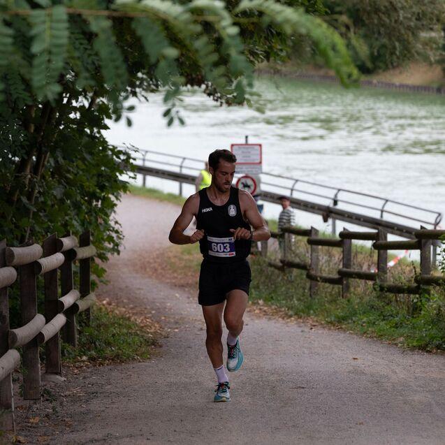 Läufer in Sportkleidung kontrolliert seine Uhr auf einem Weg entlang eines grünen Gewässers, umgeben von Bäumen.
