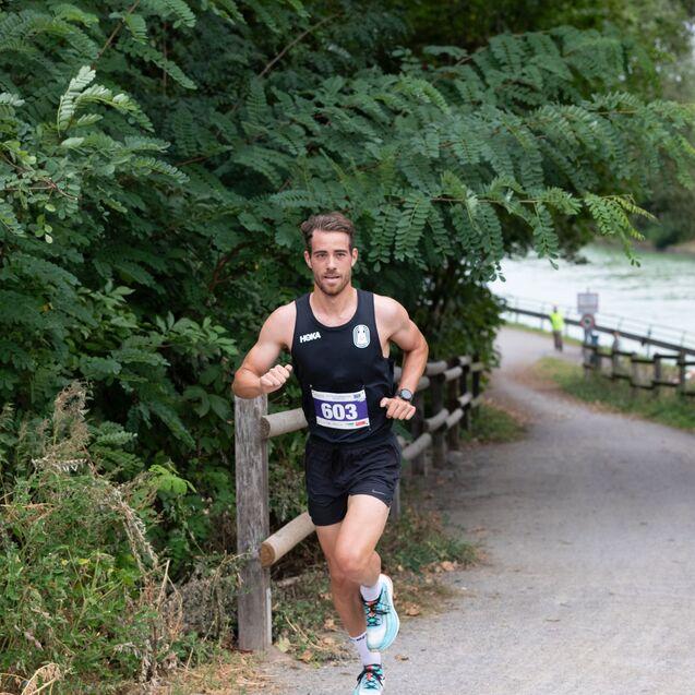 L&auml;ufer in Sportkleidung rennt auf einem schmalen Weg entlang einer gr&uuml;nen Uferlandschaft.