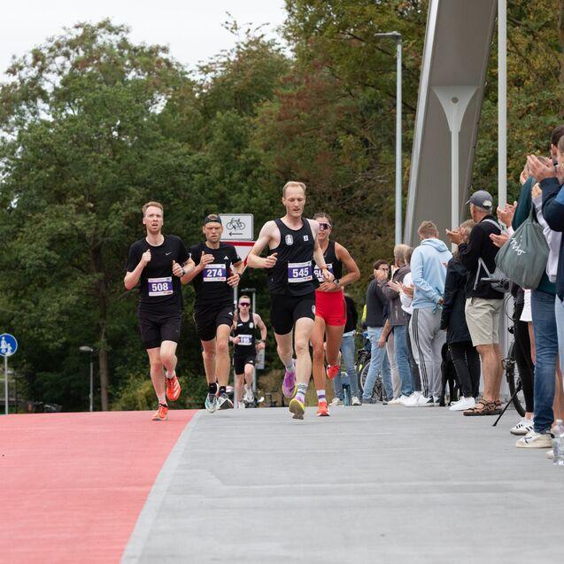 Vier L&auml;ufer auf einer roten Sportstrecke, applaudiert von Zuschauern, umgeben von Baumkulisse und urbaner Umgebung.