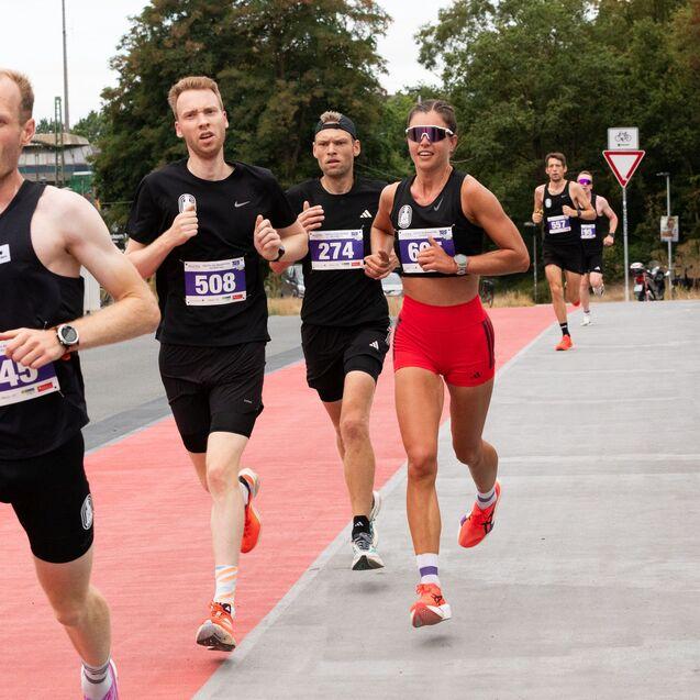 Vier L&auml;ufer in Rennkleidung laufen auf einer roten Strecke, w&auml;hrend Zuschauer am Rand applaudieren.