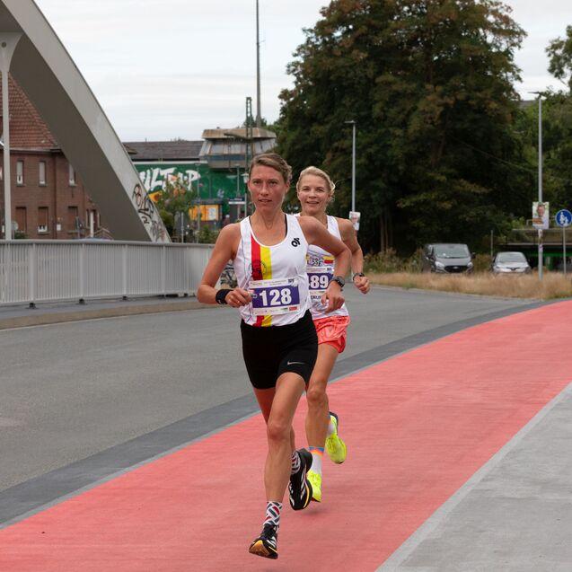 Zwei Frauen in Sportkleidung laufen auf einer roten Laufbahn, begleitet von st&auml;dtischer Umgebung und B&auml;umen im Hintergrund.