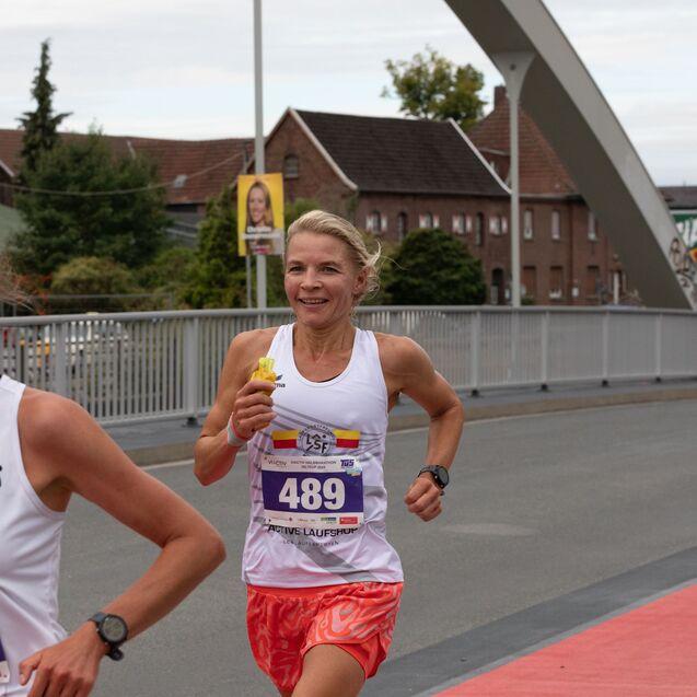 Zwei weibliche L&auml;uferinnen in Sportkleidung &uuml;berqueren eine Br&uuml;cke w&auml;hrend eines Wettlaufs. Eine h&auml;lt einen Snack in der Hand.