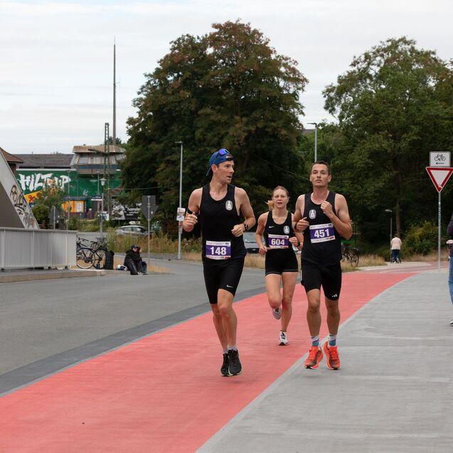 Drei L&auml;ufer in schwarz mit Startnummern auf einer roten Laufbahn, umgeben von Zuschauern und B&auml;umen.