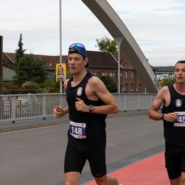 Zwei m&auml;nnliche L&auml;ufer in schwarzen Trikots mit Nummern auf der Brust joggen auf einer roten Laufstrecke.
