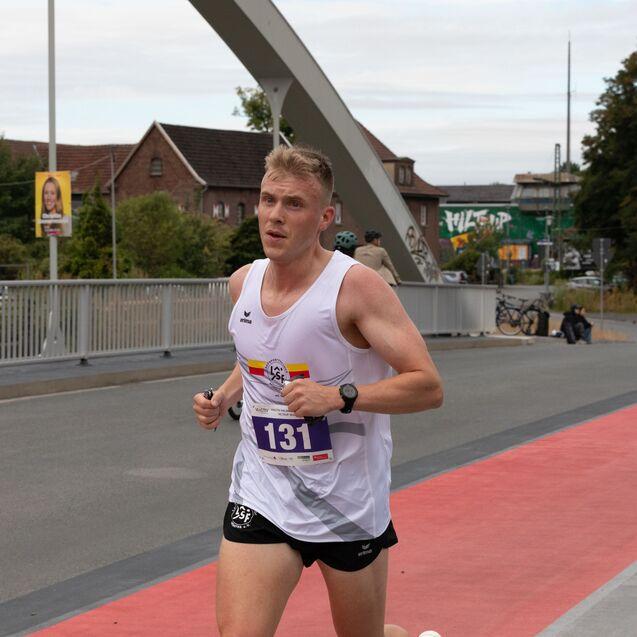 L&auml;ufer in Sportbekleidung mit Startnummer 131 auf einer roten Laufbahn, umgeben von urbaner Landschaft.
