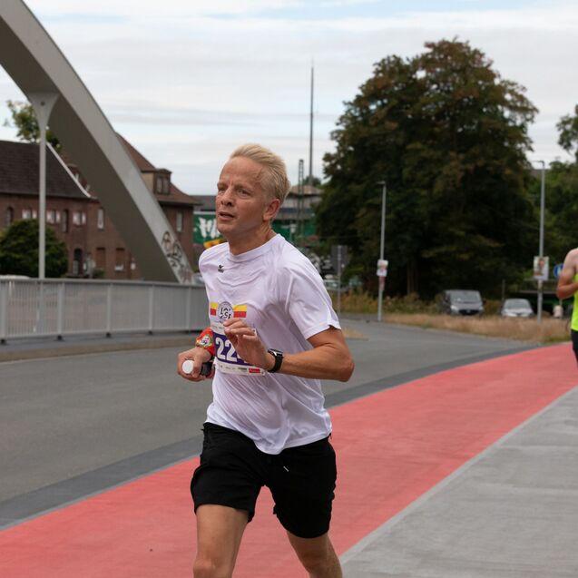 L&auml;ufer der Nummer 222 sprintet auf einer roten Laufstrecke, w&auml;hrend andere Teilnehmer im Hintergrund vorbeilaufen.