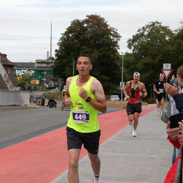 L&auml;ufer in leuchtend gelbem Shirt mit Startnummer 449, w&auml;hrend Zuschauer ihm applaudieren. Hintergrund zeigt eine Laufstrecke.