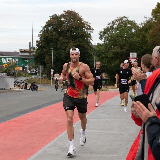 L&auml;ufer in buntem Shirt und Shorts &uuml;berquert eine Br&uuml;cke, w&auml;hrend Zuschauer ihn anfeuern. Weitere L&auml;ufer im Hintergrund.