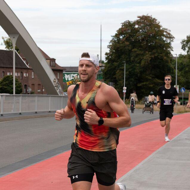 L&auml;ufer in buntem Shirt joggt auf einer roten Strecke, w&auml;hrend andere Teilnehmer im Hintergrund vorbeilaufen.