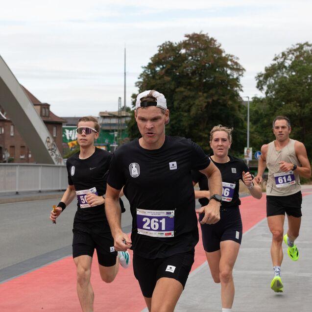 Vier aktive L&auml;ufer in schwarzen Trikots w&auml;hrend eines Wettlaufs auf einer roten Laufstrecke.