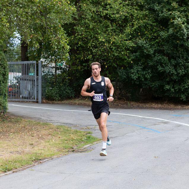 L&auml;ufer in Sportkleidung mit Startnummer 603 l&auml;uft auf einem Weg, umgeben von B&auml;umen und einem Tor im Hintergrund.