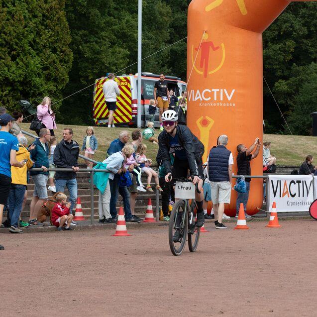 Radfahrerin mit der Nummer &bdquo;1. Frau&ldquo; f&auml;hrt in ein Veranstaltungsgel&auml;nde, umgeben von Zuschauern und Werbebannern.
