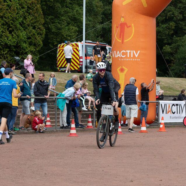 Radfahrerin mit Nummernschild &bdquo;1. Frau&ldquo; f&auml;hrt an einem Publikum und einem orangefarbenen Inflatable vorbei.