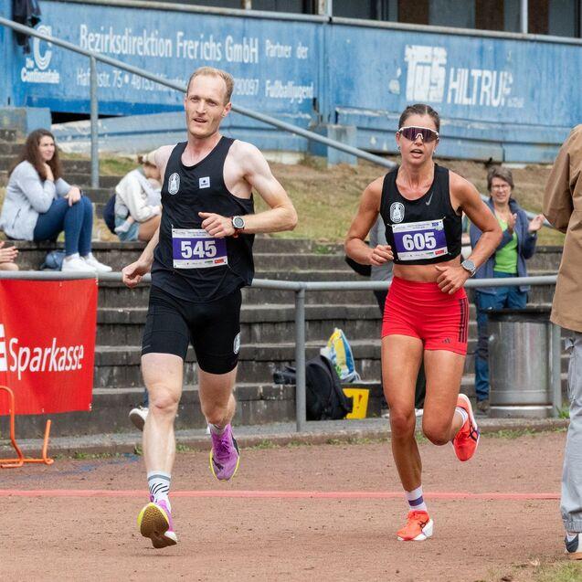 Zwei L&auml;ufer, Nummern 545 und 605, sprinten auf einer Laufbahn, w&auml;hrend Zuschauer applaudieren.