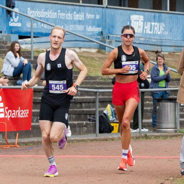 Zwei L&auml;ufer in Sportkleidung &uuml;berqueren die Ziellinie eines Wettkampfs, Zuschauer jubeln im Hintergrund.
