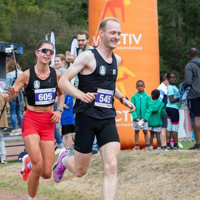 Zwei L&auml;ufer, einer in schwarzer und einer in roter Sportkleidung, sprinten am Rand einer Laufstrecke. Im Hintergrund Zuschauer.